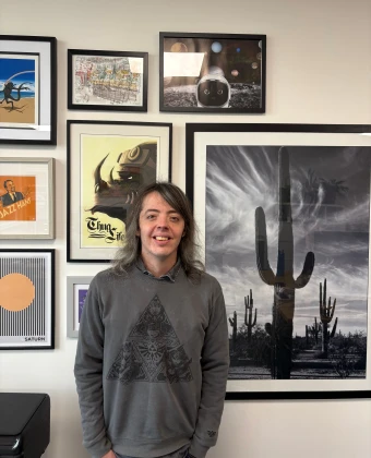 Jamie, a gentleman with shoulder length hair and a grey jumper, standing in front of a picture collage wall.