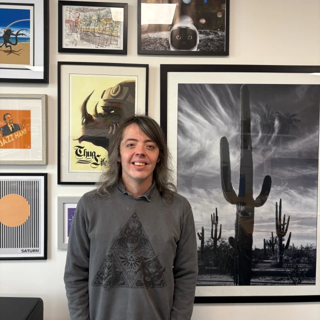 Jamie, a gentleman with shoulder length hair and a grey jumper, standing in front of a picture collage wall.