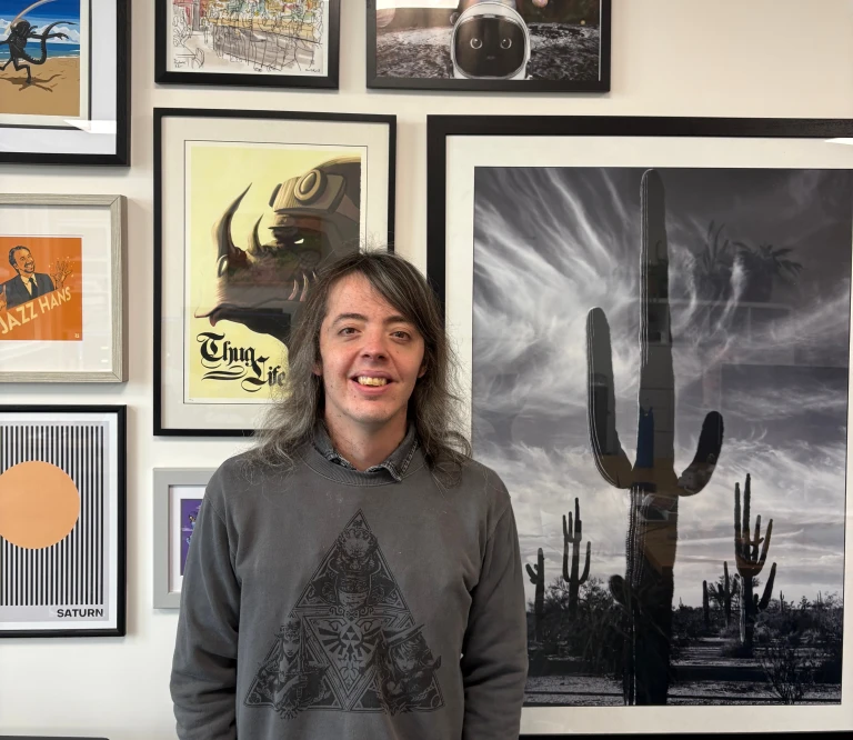 Jamie, a gentleman with shoulder length hair and a grey jumper, standing in front of a picture collage wall.